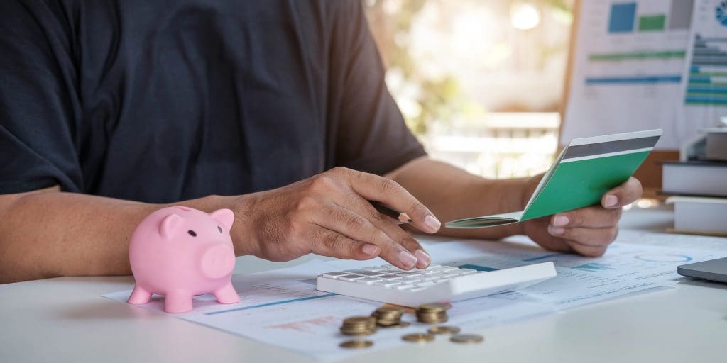 Person using calculator with piggy bank and coins on desk.  Concept of budgeting, debt consolidation. 