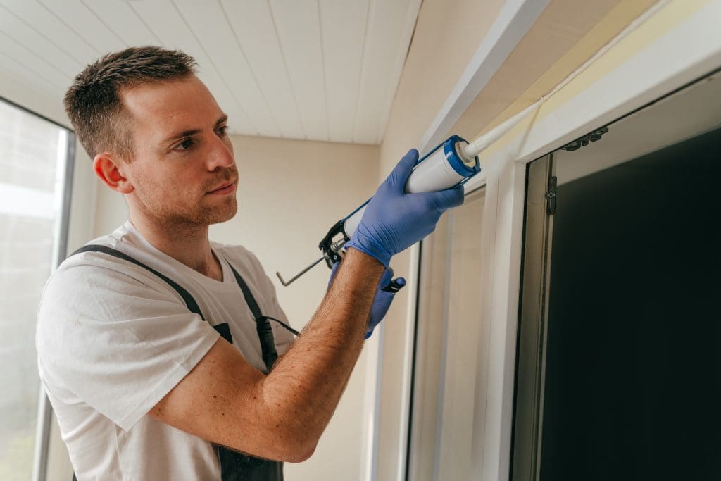 Young man wearing overalls sealing cracks between window and trim using waterproof silicone caulk on the balcony.